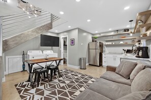 Cozinha moderna com eletrodomésticos brancos, área de jantar com mesa e cadeiras, sofá e escada que leva a um andar superior.