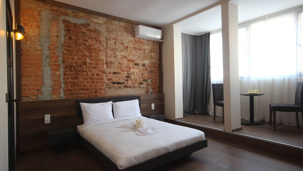 Een hotelkamer met een bakstenen muur, een bed met witte lakens, een klein tafeltje met een stoel en een groot raam met gordijnen.