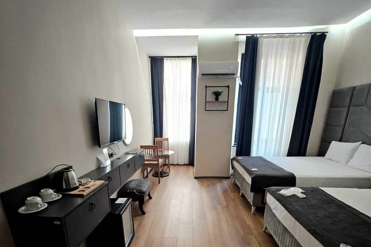 A hotel room with a bed, a desk with a computer, a chair, a TV, and a small table with cups.