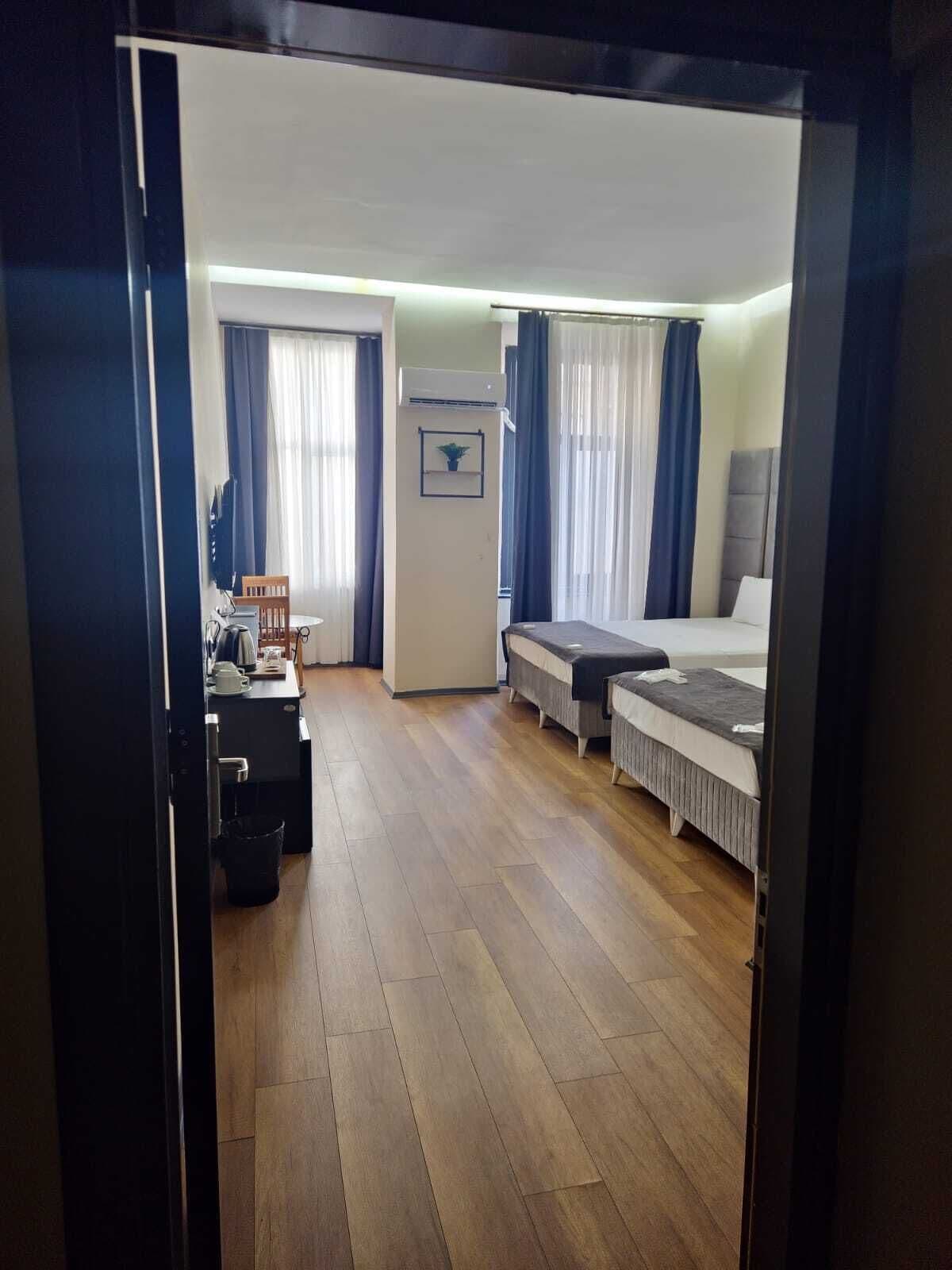 A hotel room with a wooden floor, a bed, a desk with a chair, a small table with a plant, and a TV.