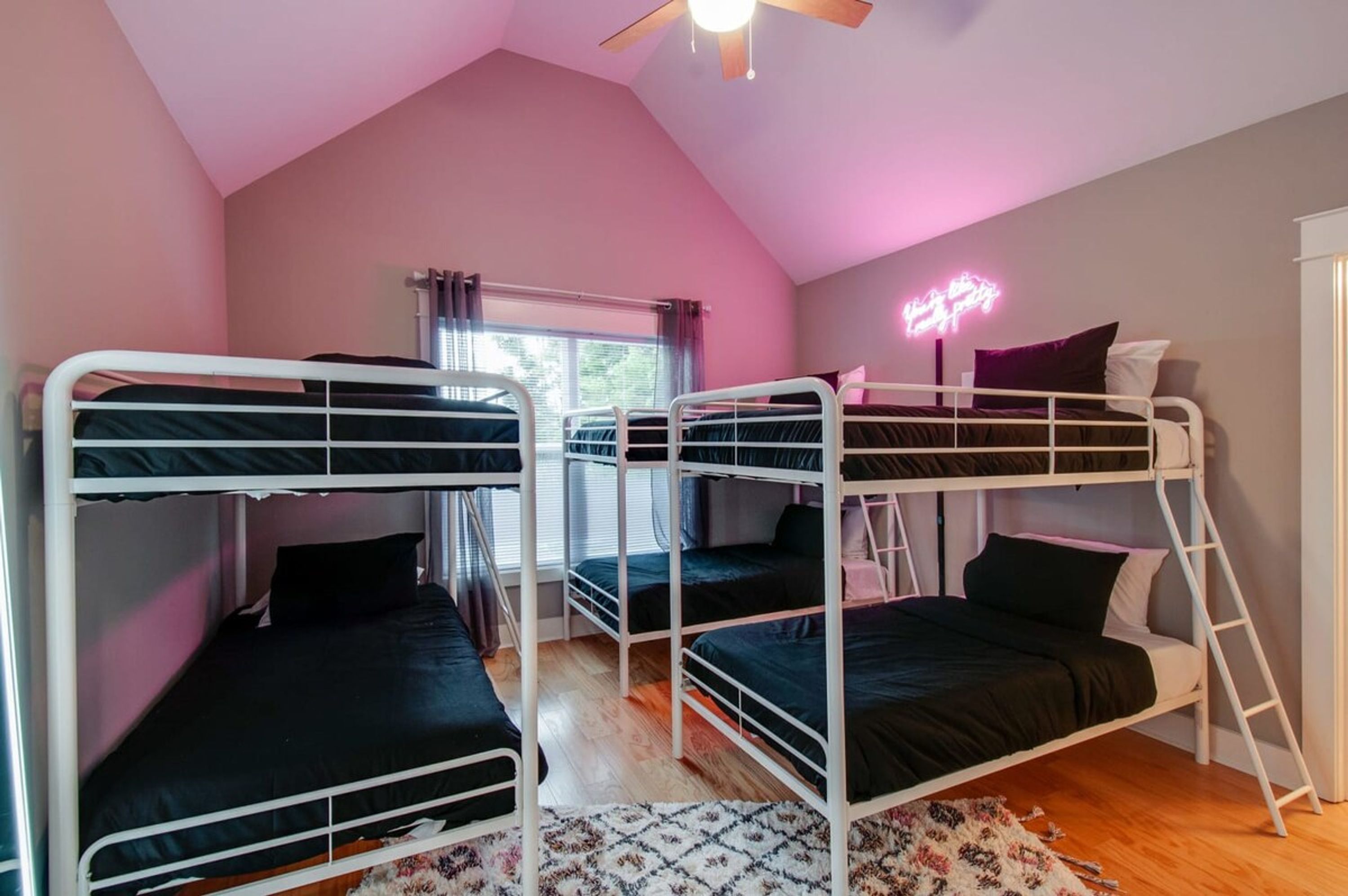 Une chambre de dortoir avec des lits superposés, un ventilateur de plafond, une fenêtre avec des rideaux et un néon sur le mur.
