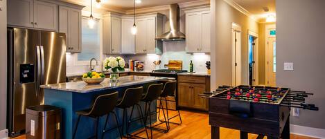 A modern kitchen with a central island, bar stools, a stainless steel refrigerator, and a foosball table.