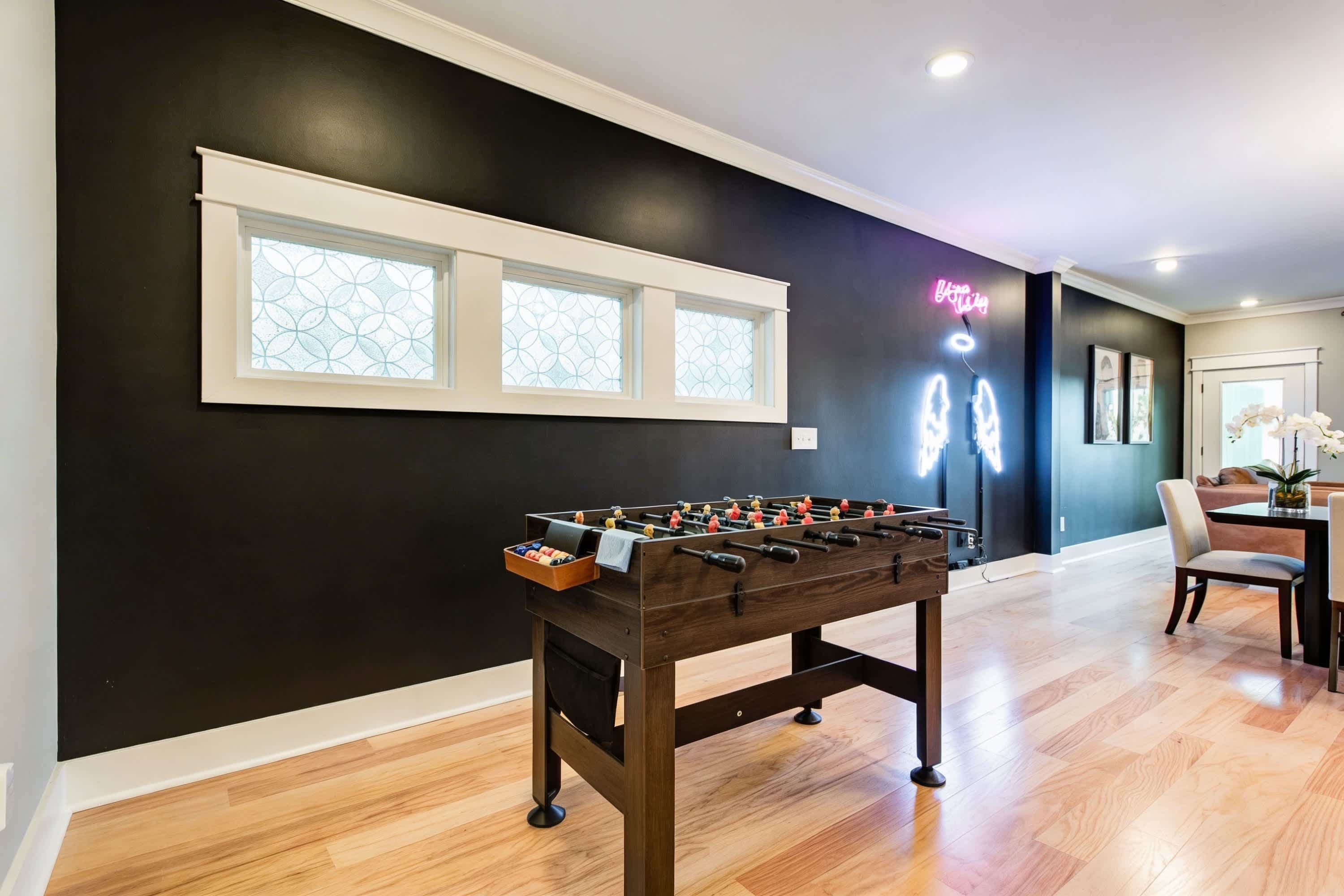 Une pièce avec une table de baby-foot, une fenêtre ornée d’un motif géométrique et un panneau lumineux au néon sur le mur.