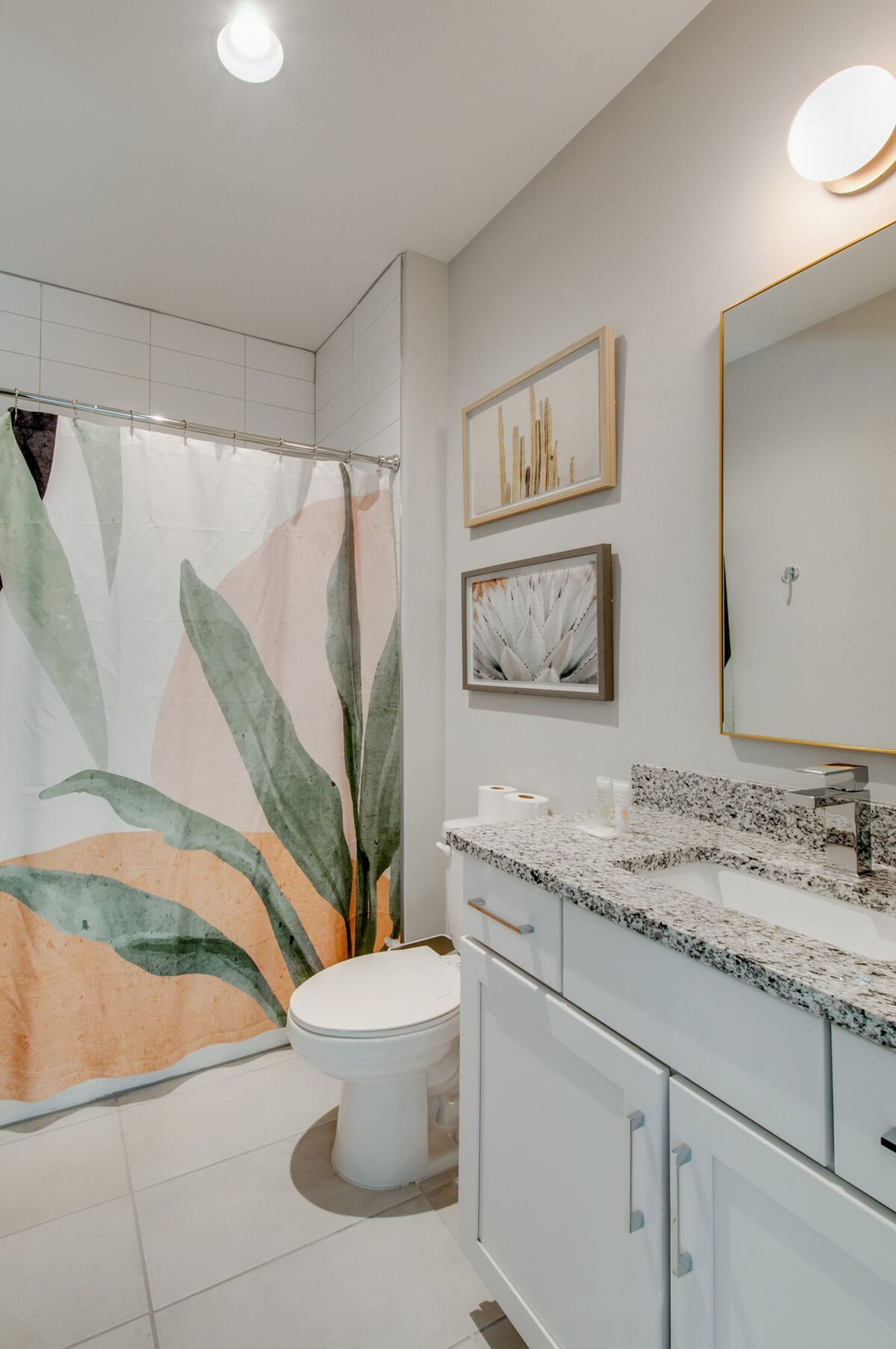 Un baño moderno con cortina de ducha estampada con hojas tropicales, un inodoro, un lavamanos con encimera de granito y un espejo con marco dorado.