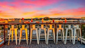 A rooftop bar with white metal chairs and tables, overlooking a cityscape with buildings and a sunset.
