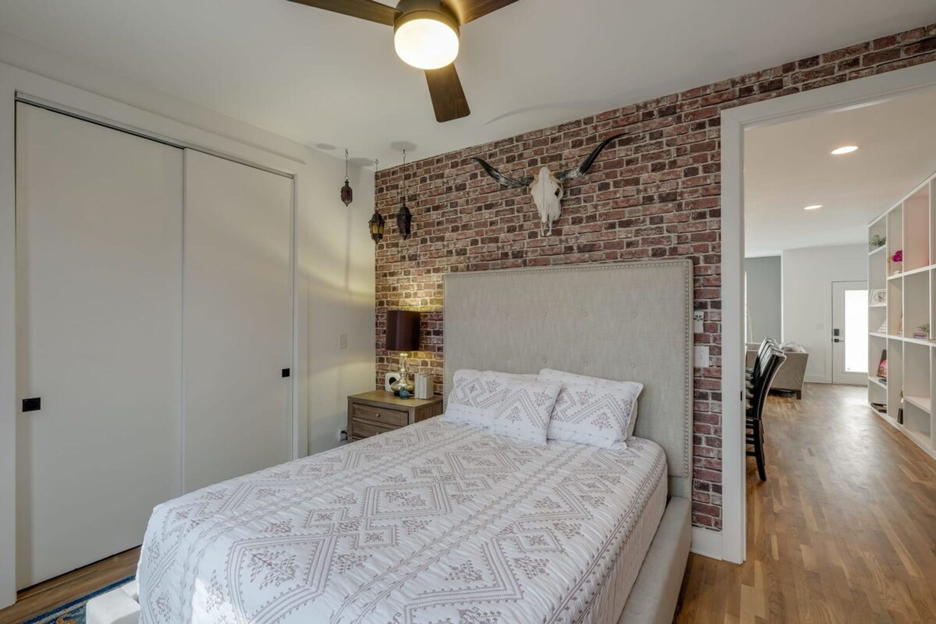 A bedroom with a brick wall, a bed with patterned bedding, a nightstand, and a ceiling fan.