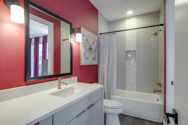 A modern bathroom with a white sink, gold faucets, and a large mirror. There is a shower with a glass door and a white toilet.