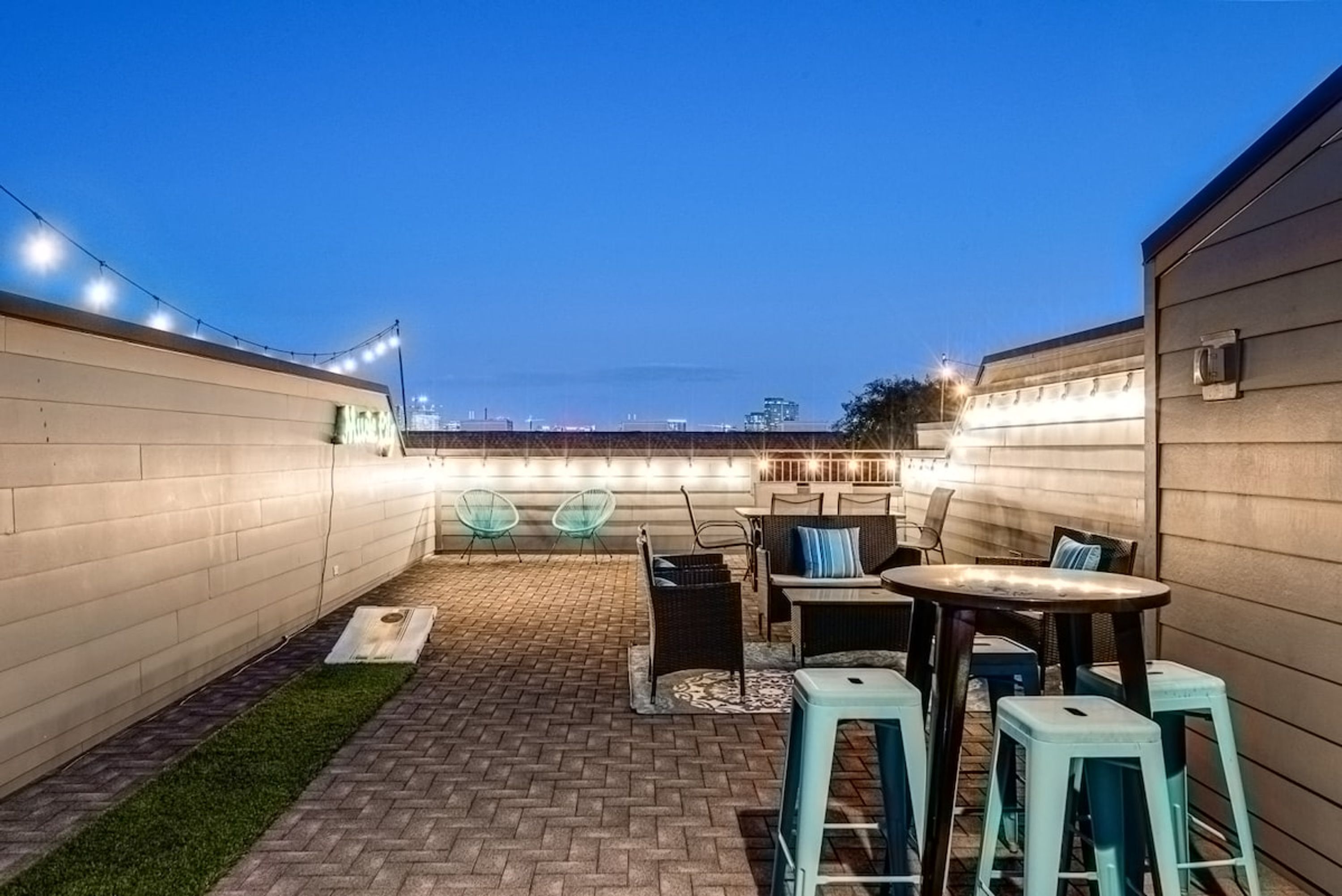 Un patio al aire libre con piso de ladrillo, césped artificial verde y mobiliario exterior, incluyendo mesas y sillas.