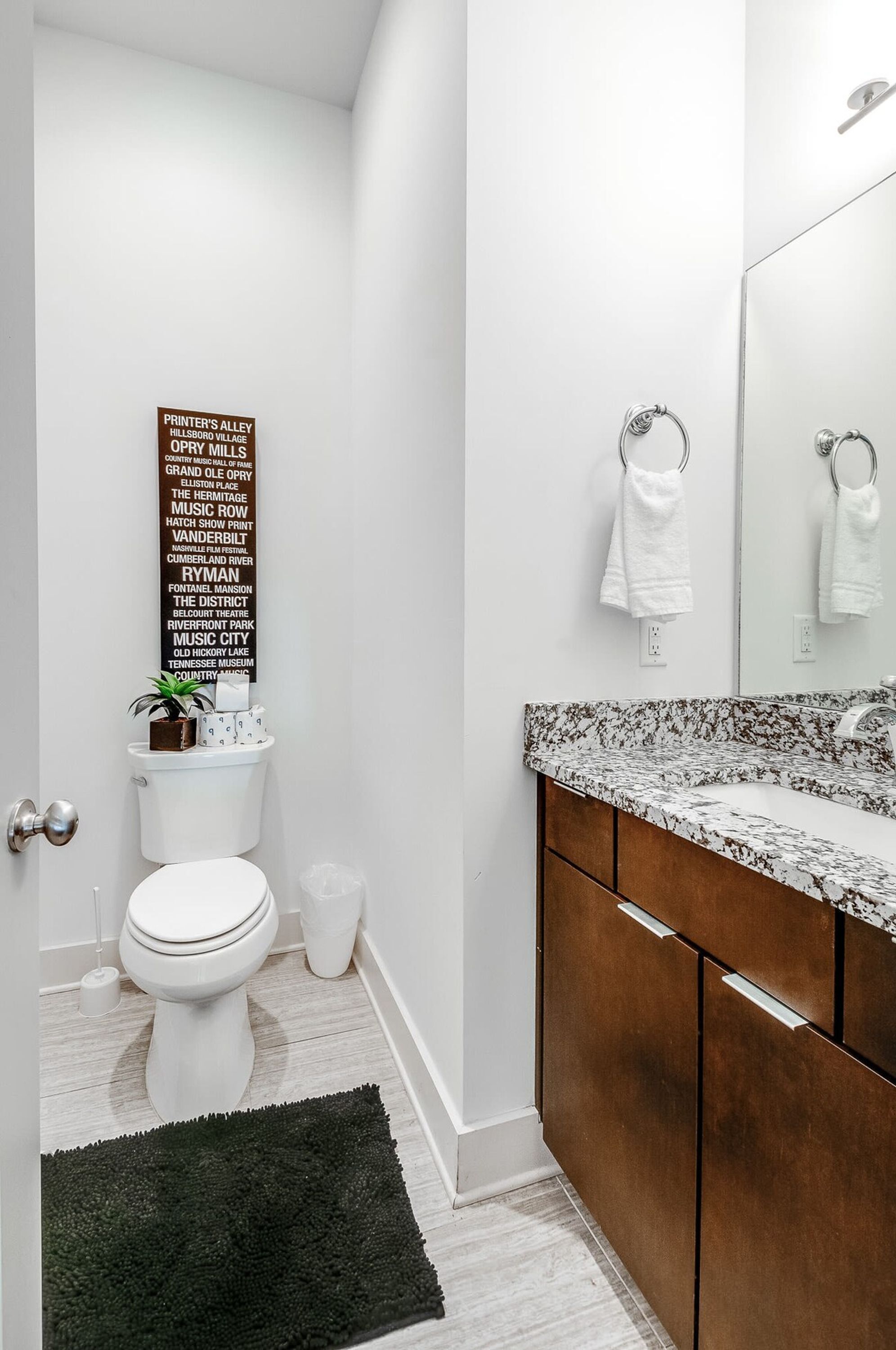 A modern bathroom with a white toilet, a black rug, a wooden vanity with a marble countertop, and a wall-mounted mirror.