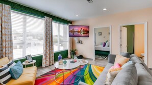 Sala de estar moderna com sofá, mesa de centro e televisão. Há uma janela com persianas e uma parede verde como destaque.