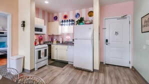Cozinha compacta com eletrodomésticos brancos, micro-ondas e geladeira. Há um pequeno espaço de refeições com uma cadeira e uma mesa. As paredes estão pintadas em tom claro e há pratos decorativos na parede.