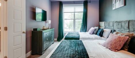 Une chambre à coucher avec un lit, une commode, un téléviseur et une fenêtre avec des rideaux.