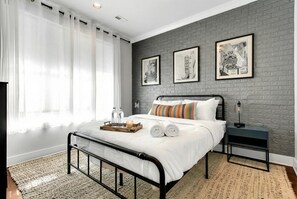 Une chambre à coucher moderne avec un lit en métal, une table de chevet et une commode. Trois tableaux encadrés ornent le mur, il y a une fenêtre avec des rideaux et un tapis sur le sol.