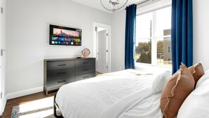 A modern bedroom with a large bed, a flat-screen TV mounted on the wall, a dresser, and a view of the outside through a large window with blue curtains. - Near Gulch 12 South Book Luxury Now (Nashville)