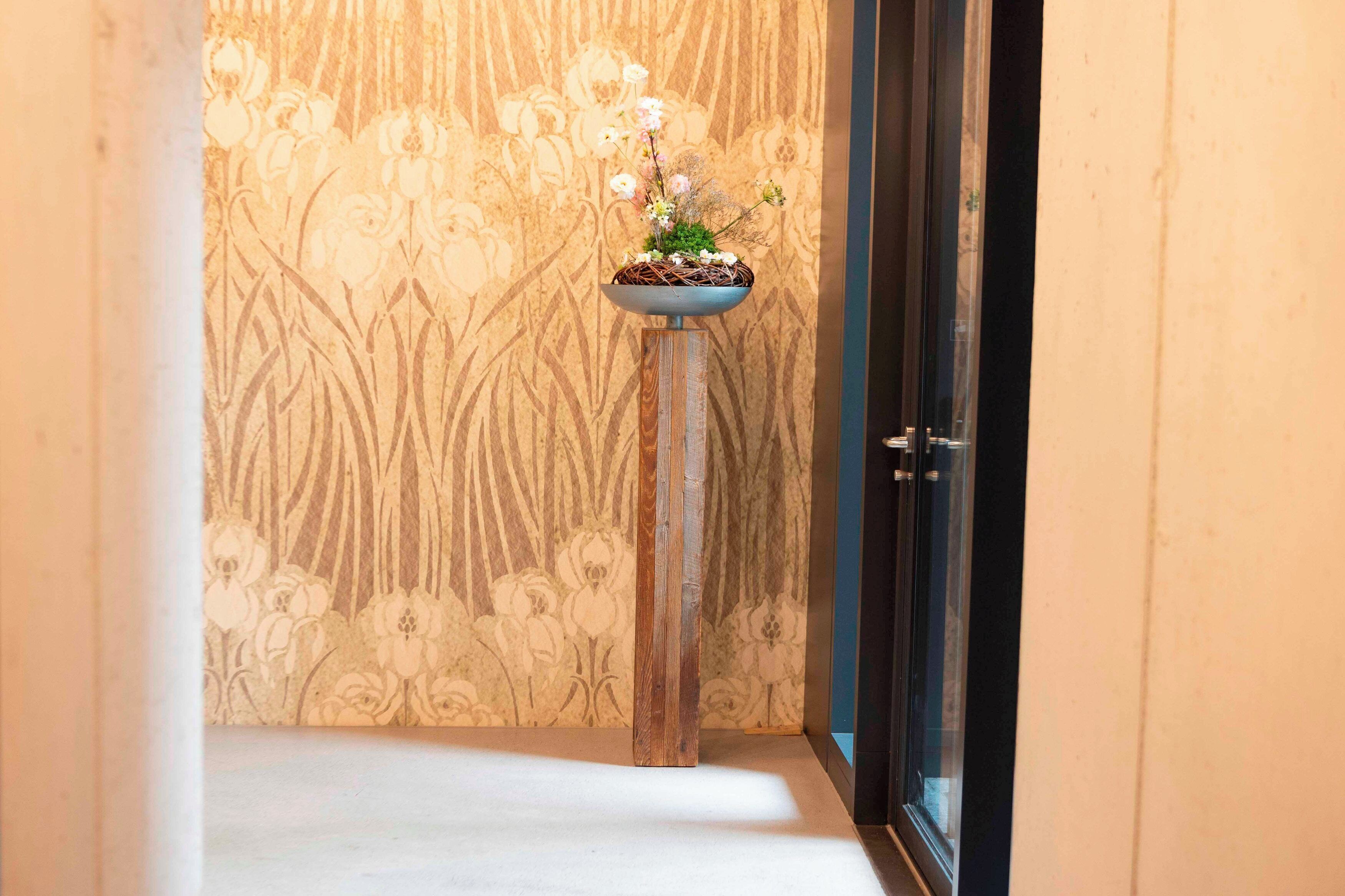 Ein schmaler Flur mit floralem Wandpapier und Holzboden. Gegen die Wand ist ein kleiner Holzschemel mit einer blauen Schüssel voller Blumen gestellt.