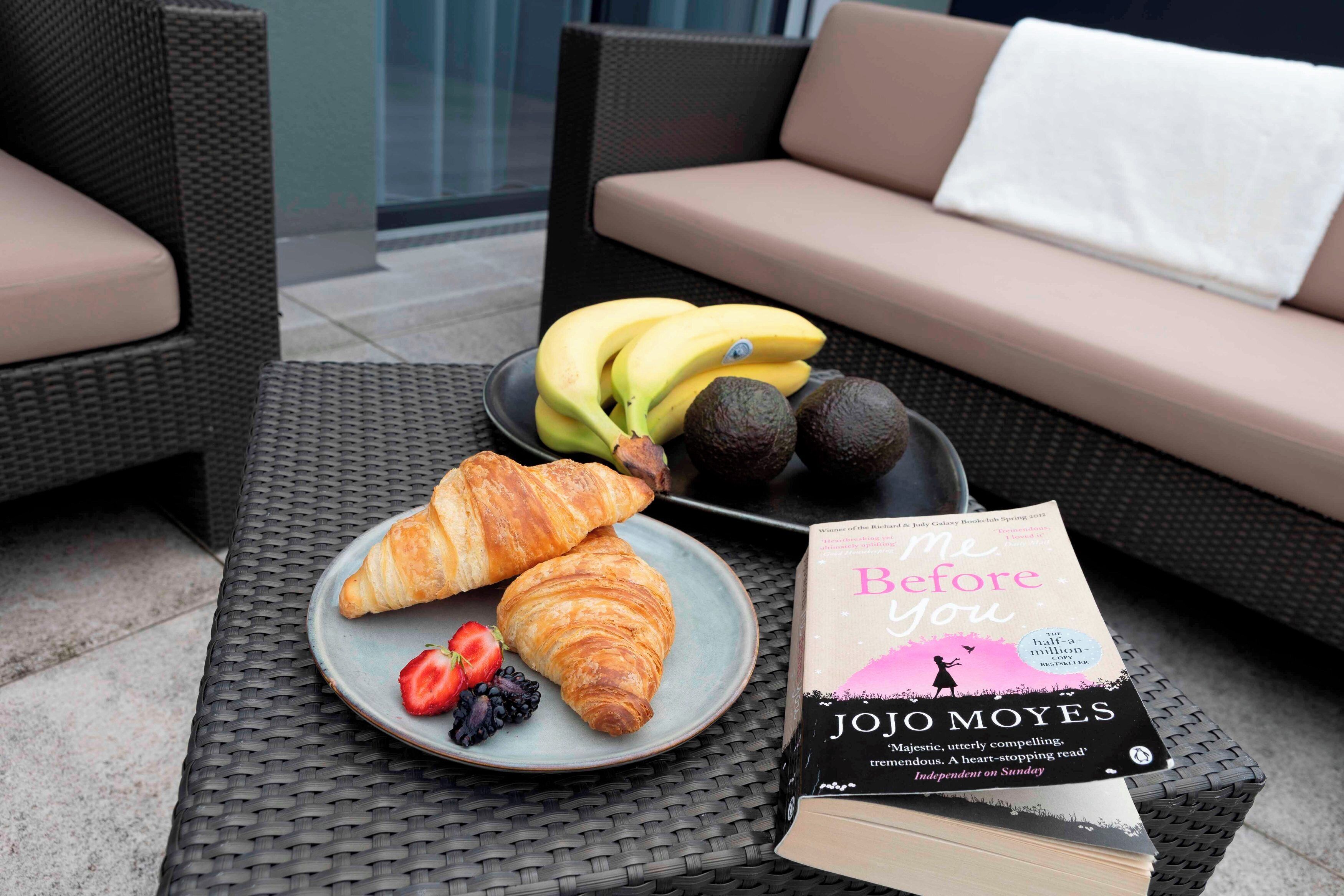 Une table en osier sur laquelle se trouvent une assiette de croissants, des fraises, des mûres et un livre intitulé « Avant toi » de Jojo Moyes.