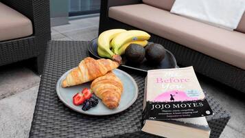 Une table en osier sur laquelle se trouvent une assiette de croissants, des fraises, des mûres et un livre intitulé « Avant toi » de Jojo Moyes.