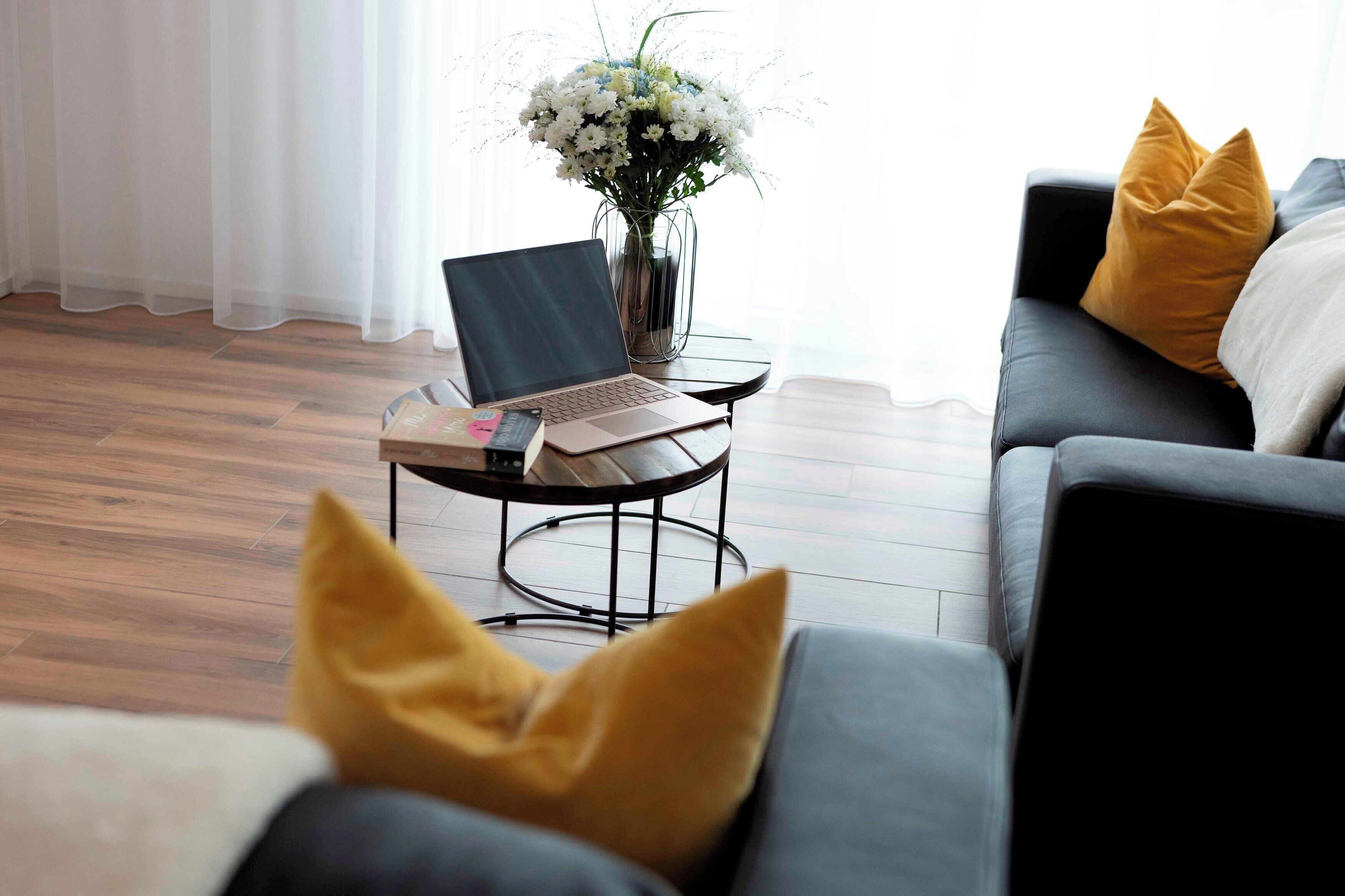 A modern living room with a laptop on a small round table, a vase with flowers, and a sofa with cushions.