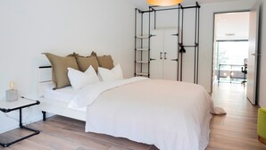 A modern bedroom with a white bed, a black pendant light, and a wooden floor. - THE CITY ZUG APARTMENTS (Zug)