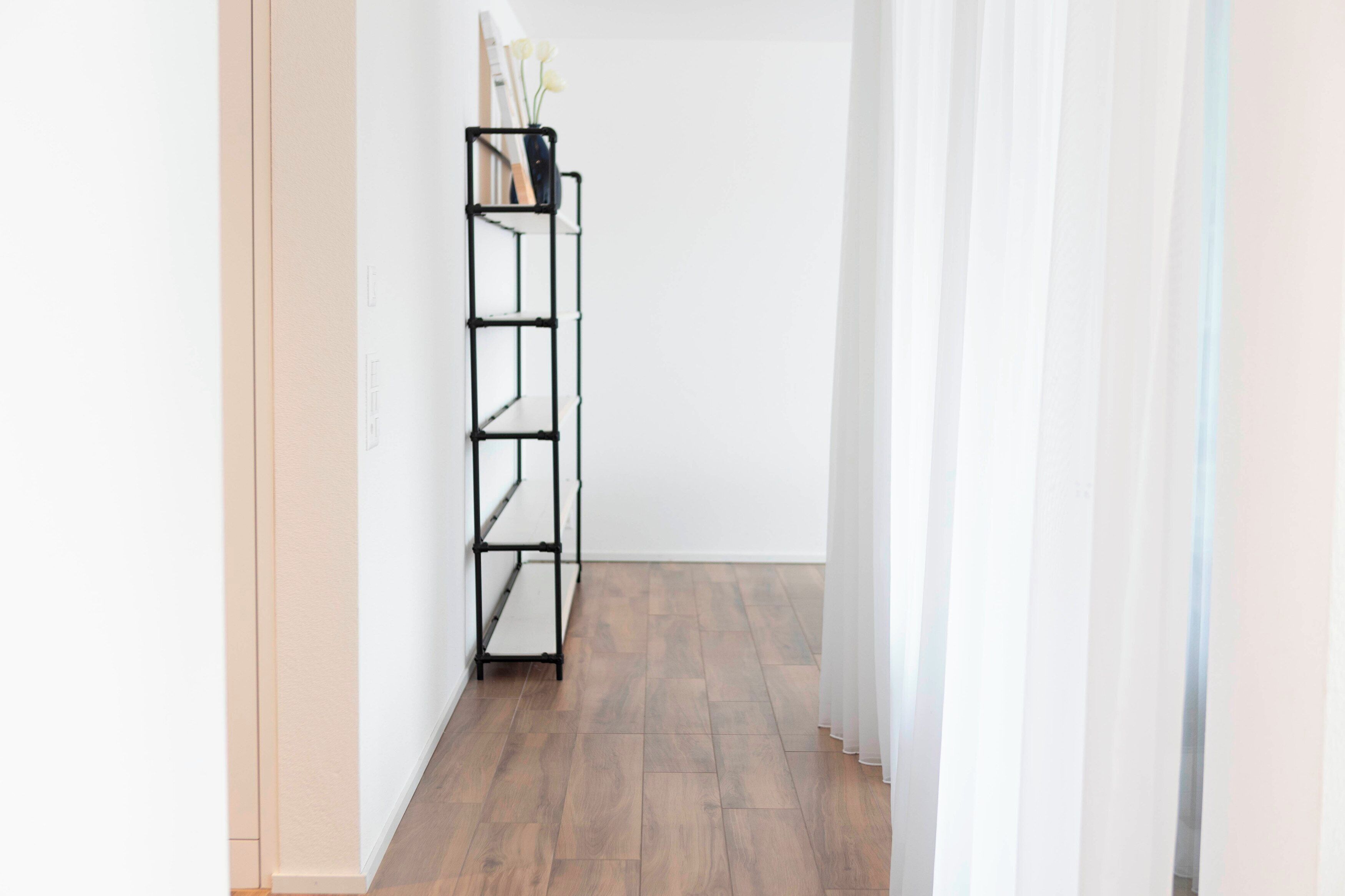 A hallway with a black metal shelf unit against a white wall, wooden flooring, and sheer white curtains.