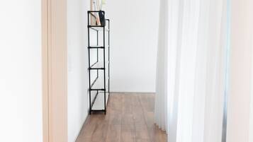 A hallway with a black metal shelf unit against a white wall, wooden flooring, and sheer white curtains.