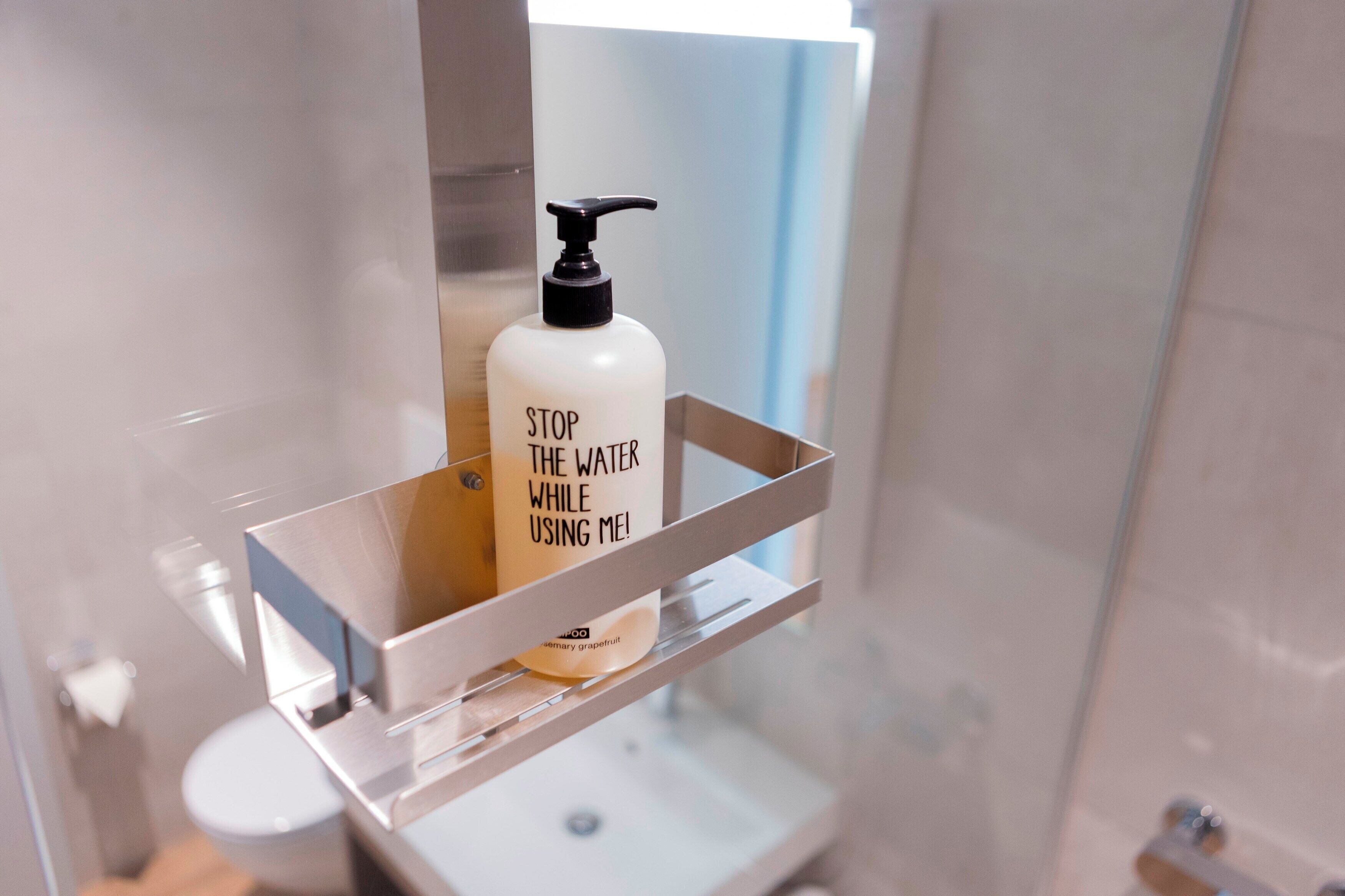 A bathroom with a white bottle of shampoo on a shelf and a mirror above it.