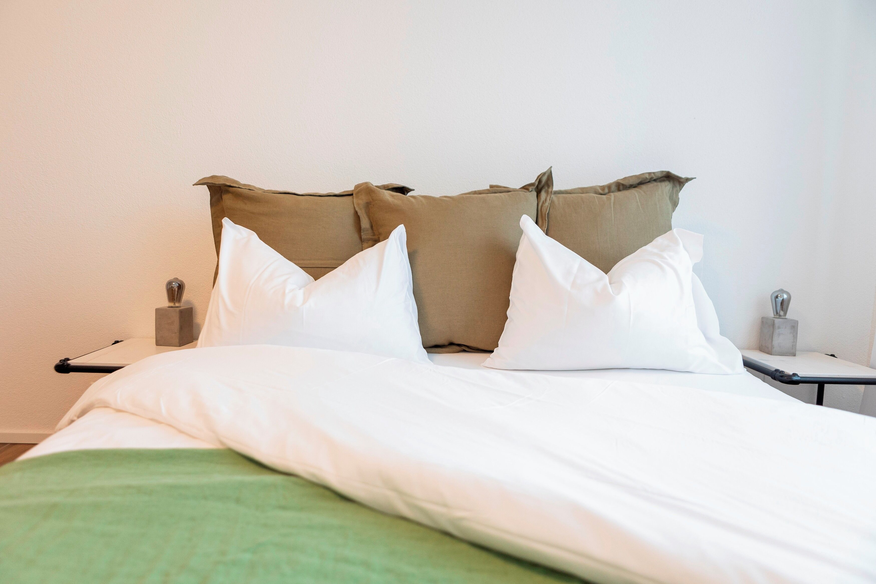 A neatly made bed with white and beige pillows, a green blanket, and a bedside table with a lamp and a small decorative item.