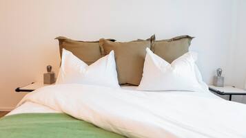 A neatly made bed with white and beige pillows, a green blanket, and a bedside table with a lamp and a small decorative item.