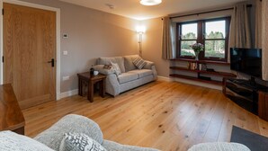 Ein gemütliches Wohnzimmer mit Holzboden, einem grauen Sofa, einem Couchtisch, einem Fernseher und einem Fenster mit Vorhängen.