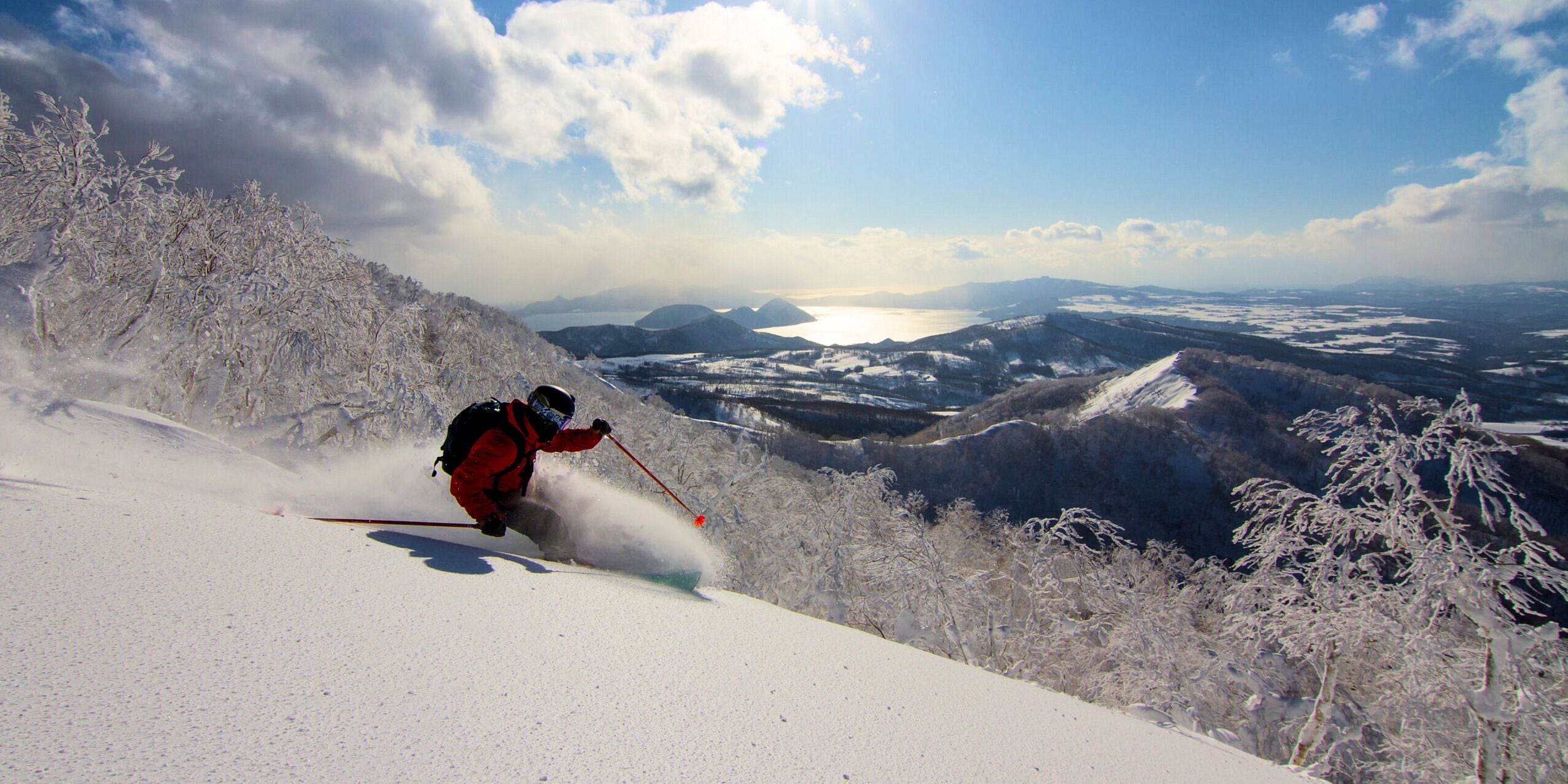 A skier on a snow-covered slope with a panoramic view of mountains and a lake in the distance.