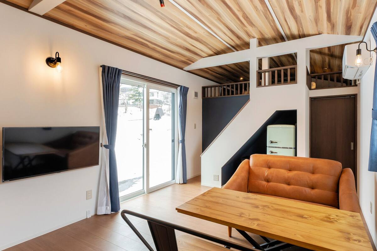 A modern interior with a wooden ceiling, a flat-screen TV mounted on the wall, a dining table with a chair, and a staircase leading to an upper level.