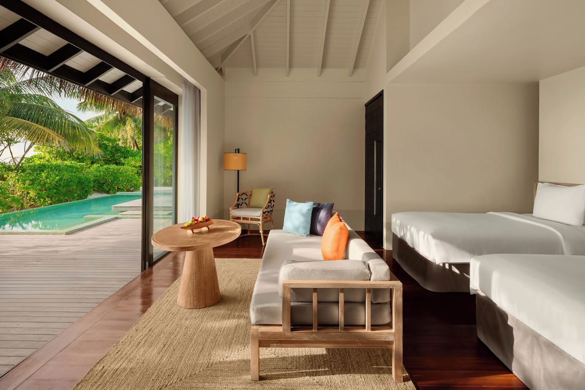 A room with a large glass door leading to a pool, a sofa with cushions, a small round table with a lamp, and a bed.