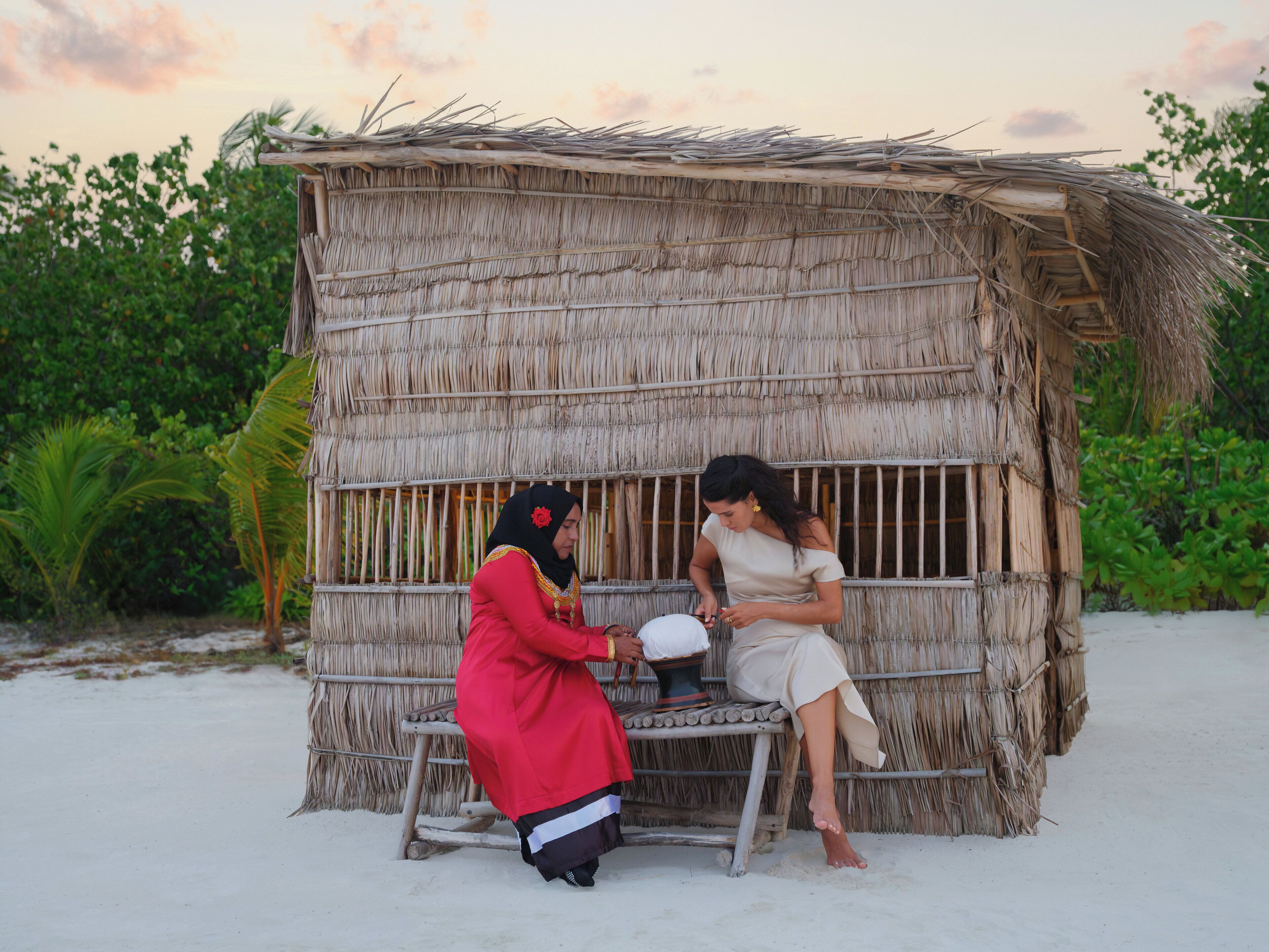 A rustic hut with a thatched roof, situated on a sandy beach with lush greenery in the background.