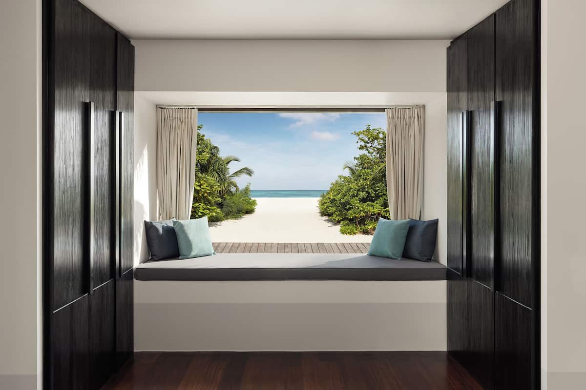 A room with a view of a beach through a large window, a built-in bench with cushions, and dark wooden walls.
