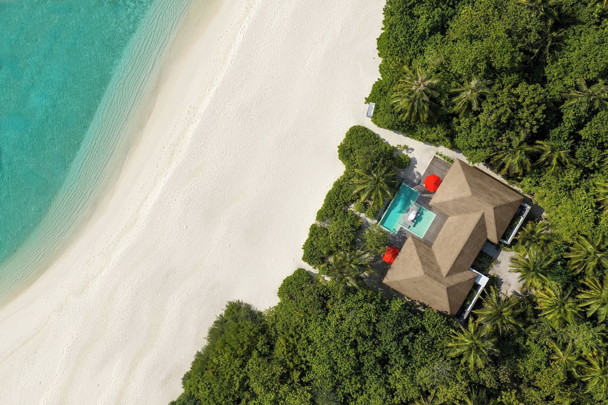 An aerial view of a beach resort with a swimming pool, palm trees, and a white sandy beach.
