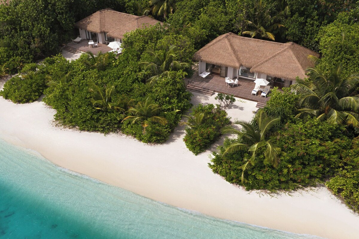 An aerial view of a beachside resort with two thatched-roof cottages surrounded by lush greenery and a sandy beach.