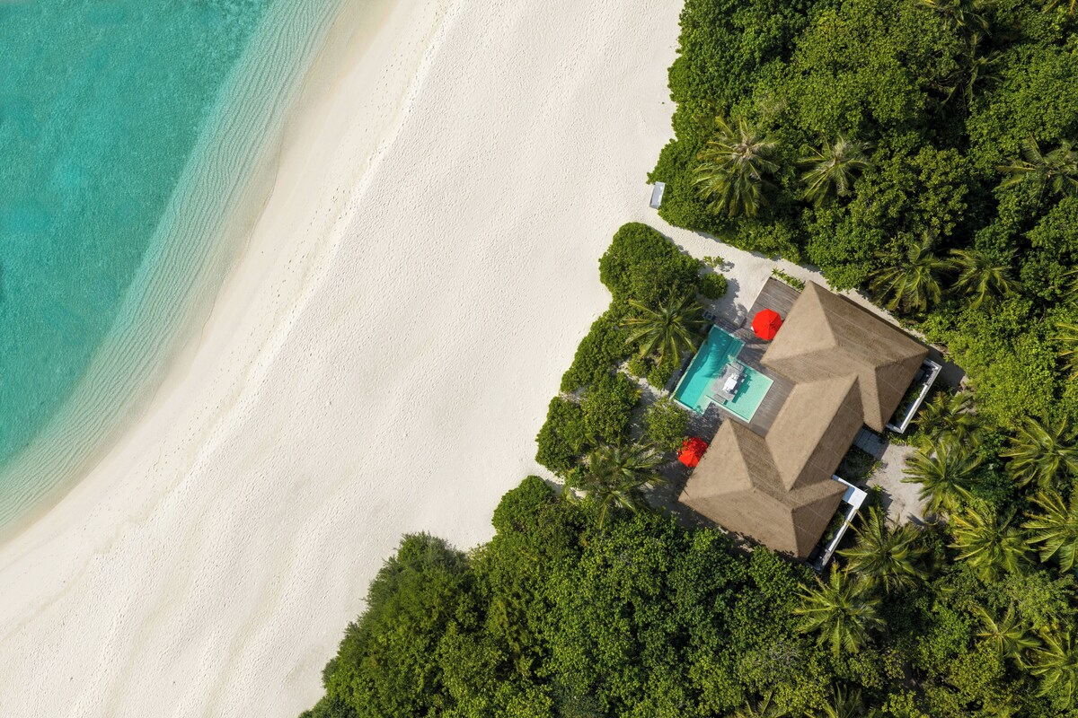 An aerial view of a beach resort with a swimming pool, palm trees, and a white sandy beach.