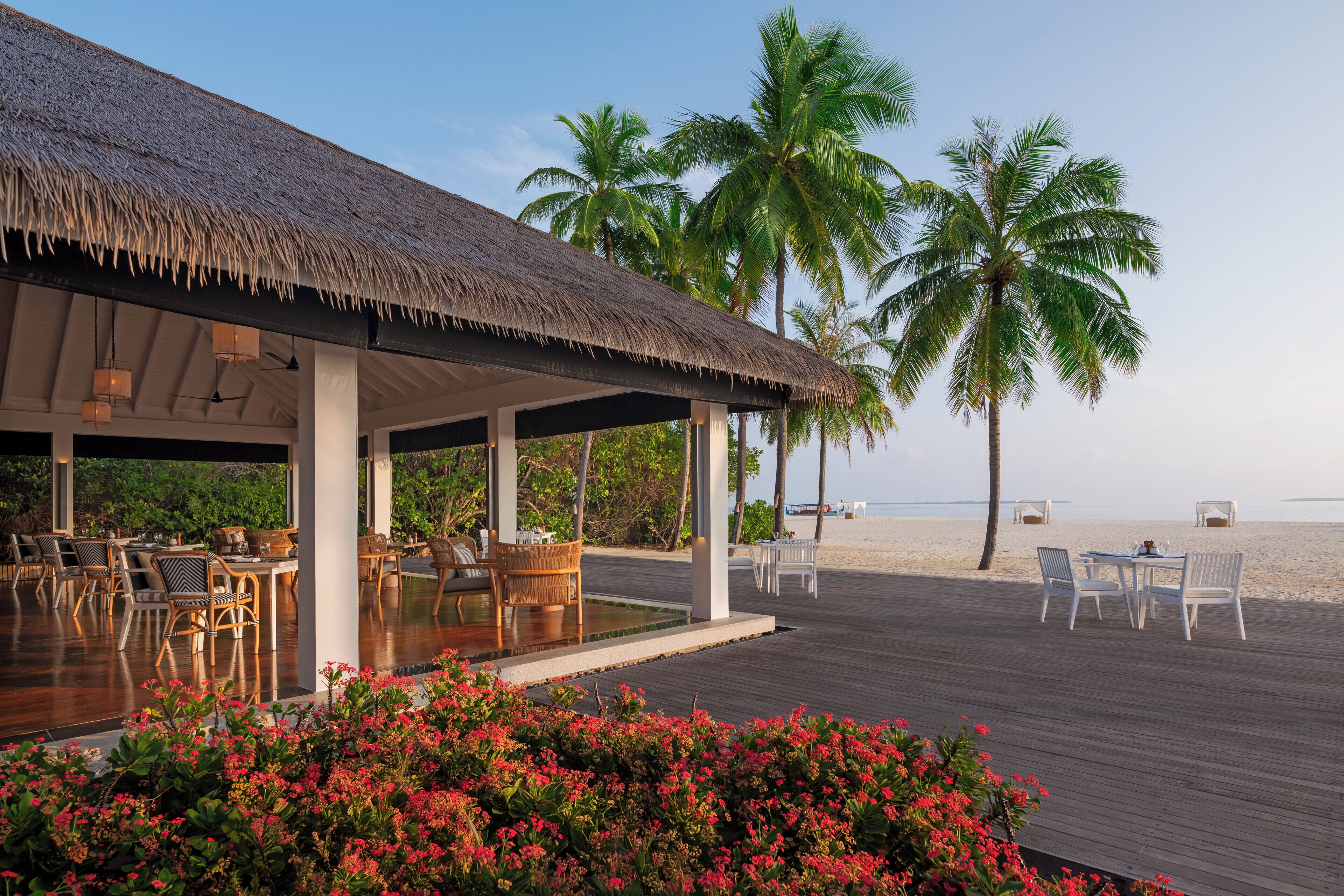 Un espacio al aire libre cubierto con mesas y sillas, una piscina y vistas a la playa y palmeras.