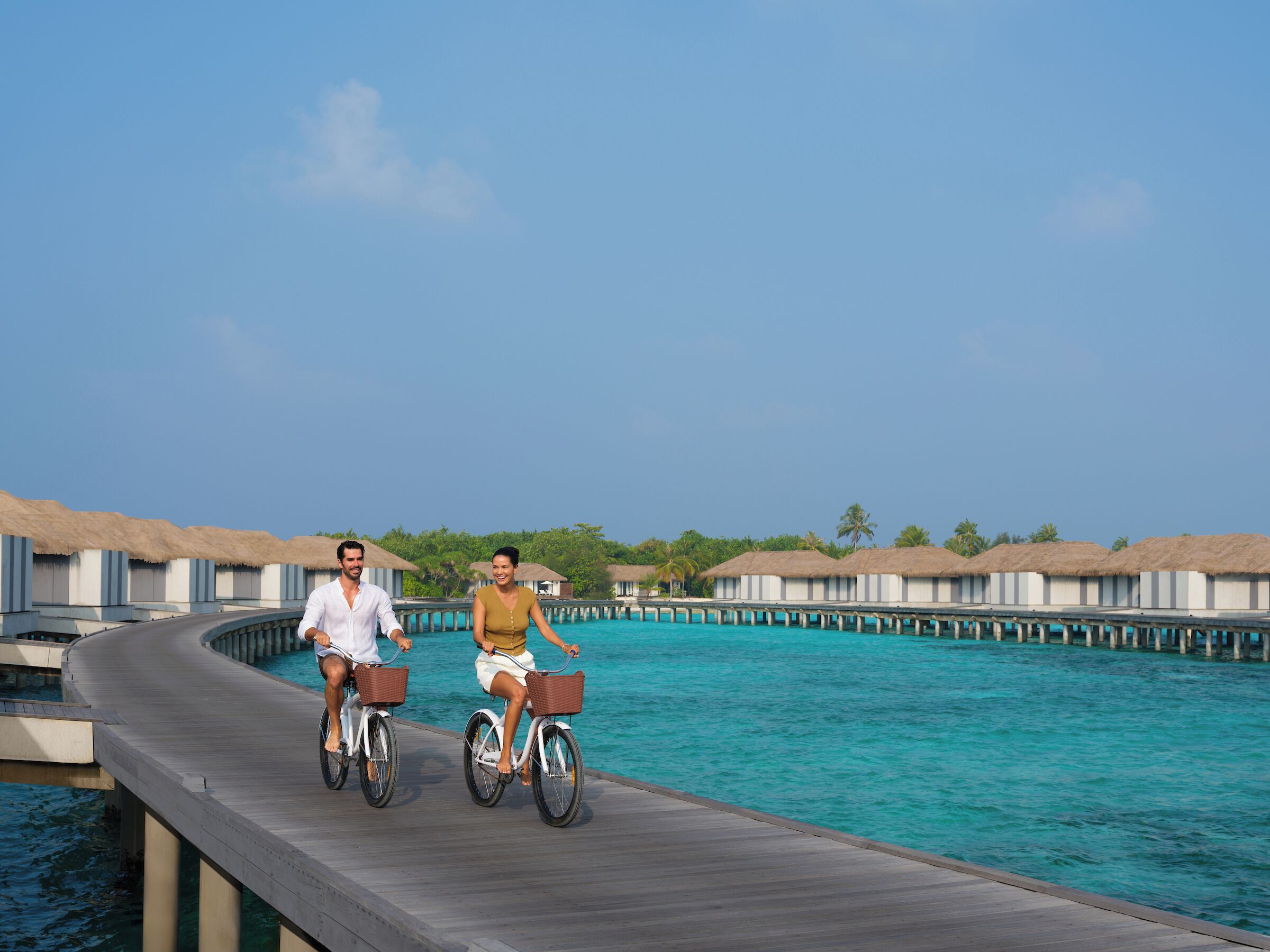 Twee personen fietsen over een houten looppad boven het water, met drijvende bungalows op de achtergrond.
