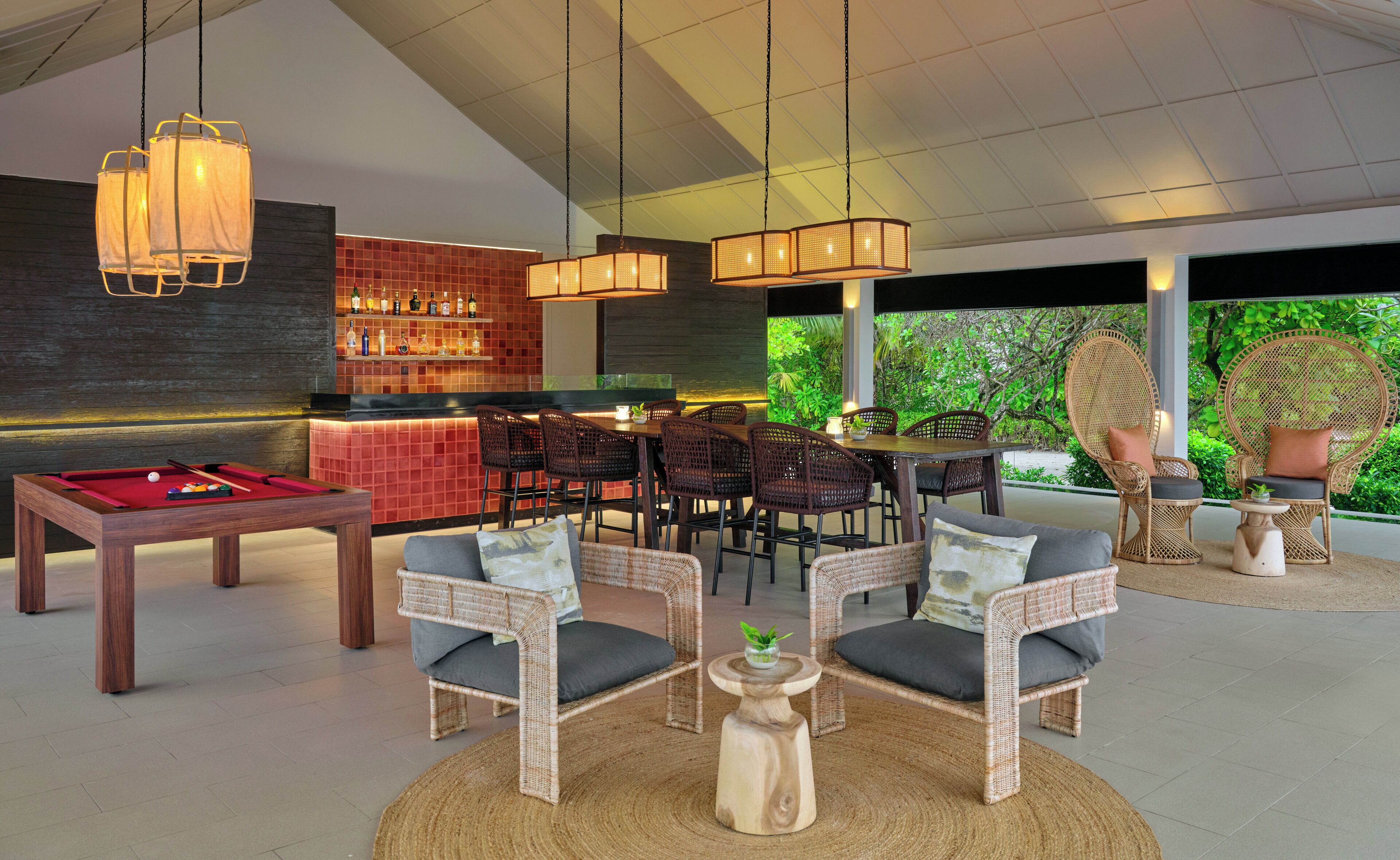 A modern interior with a pool table, bar area, wicker furniture, and a view of greenery.