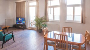 A living room with a wooden dining table, chairs, a television, a potted plant, and large windows with curtains. - Spacious 2-bedroom 2 story loft near Opera/Andrassy with AC, WiFi (Budapest)