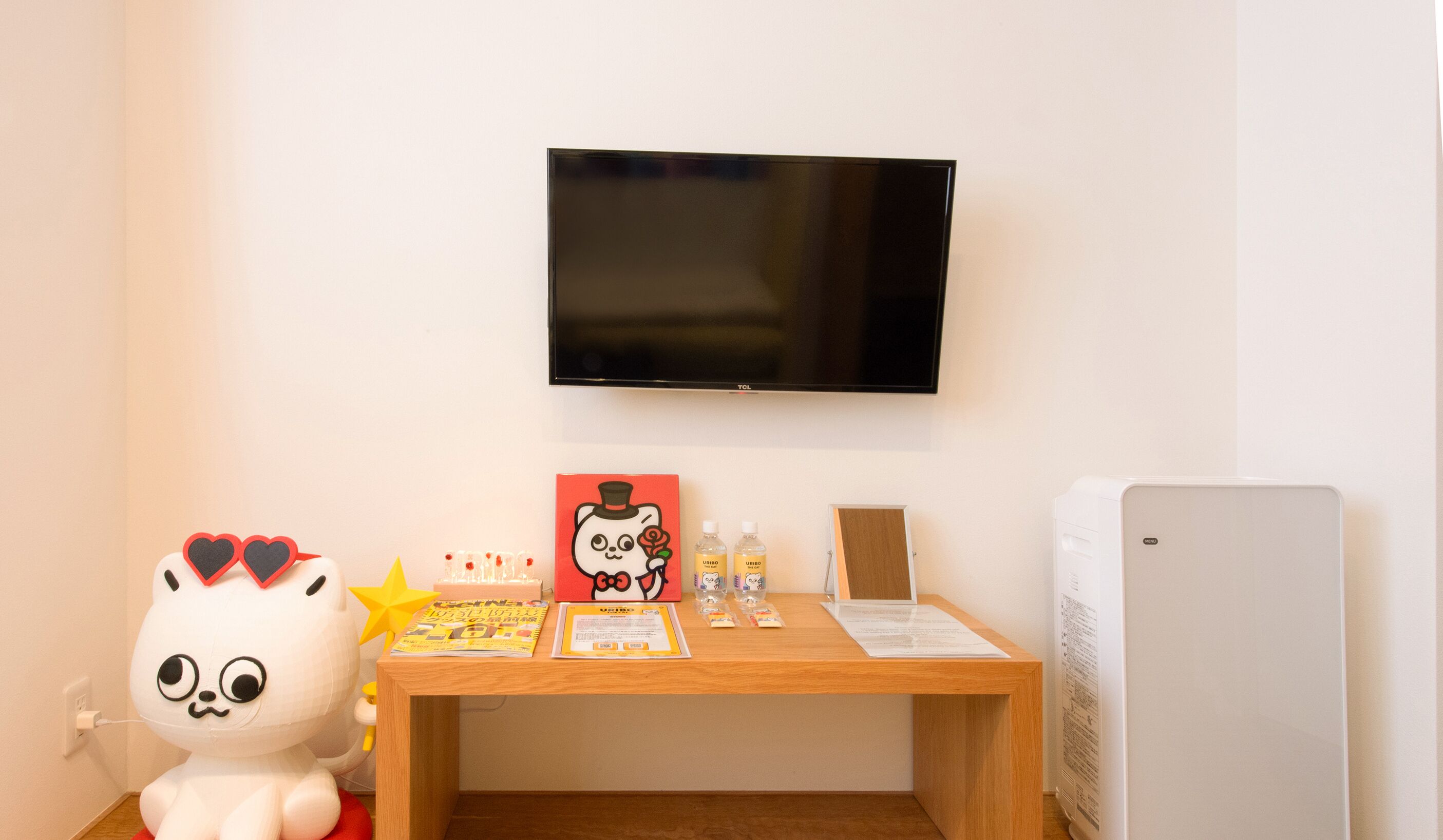 Ein Zimmer mit an der Wand befestigtem Fernseher, einem Holztisch mit einer Zeitschrift, einem Plüschtier und einem Kühlschrank.