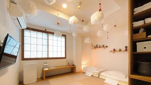 A room with tatami flooring, a futon bed, a TV on a stand, a heater, a window with a view, and a shelf with decorative items. - Fushiginayado (Kyoto)