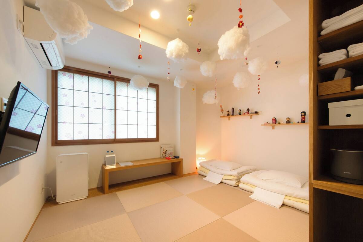 Ein Zimmer mit Tatami-Boden, einem Futon-Bett, einem Fernseher auf einem Schrank, einer Heizung, einem Fenster mit Aussicht und einem Regal mit Dekorationen.
