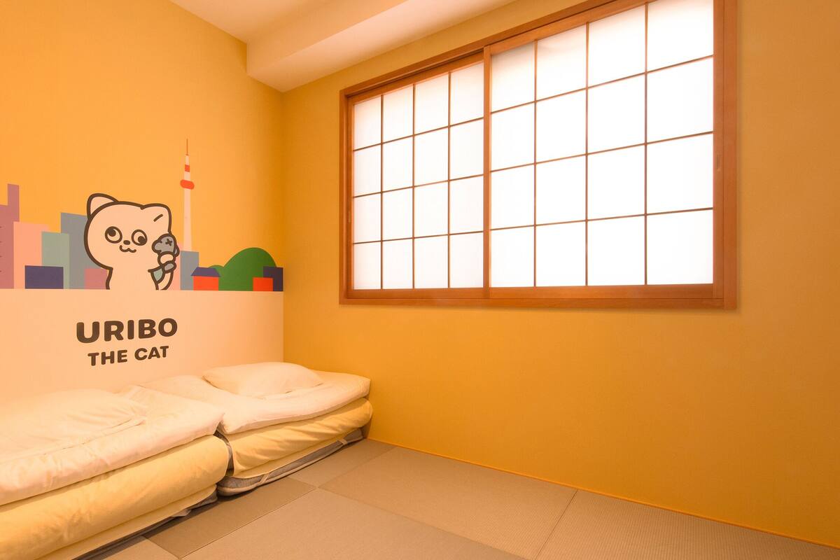 Ein Zimmer mit Futon, einem großen Fenster mit Gittermuster und einer Wand mit einer Cartoon-Katzen-Illustration und dem Text "URIBO THE CAT".