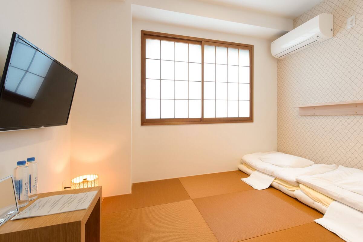 Ein Hotelzimmer mit Tatami-Boden, einem Bett, einem Fernseher und einem Fenster mit Gittermuster.