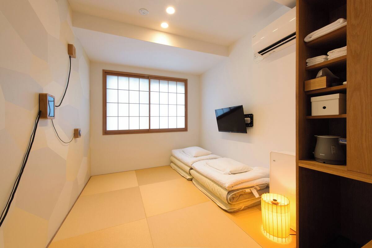 Ein kleiner, moderner Raum mit Tatami-Boden, einem Futon-Bett, einem an der Wand montierten Fernseher und einem Regal mit Handtüchern und einem Wasserkocher.