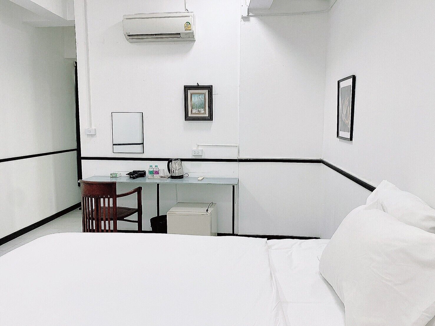 A hotel room with a bed, a desk with a chair, an air conditioner, and two framed pictures on the wall.