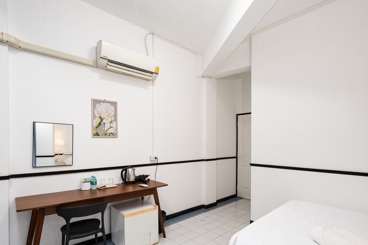 Habitación de hotel con cama, un escritorio con silla, una mesita con tetera, un espejo y un equipo de aire acondicionado.