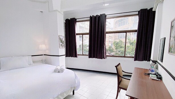 Une chambre d’hôtel avec un lit, un bureau et une chaise. Il y a des fenêtres avec des rideaux, une lampe et une plante accrochée au mur.